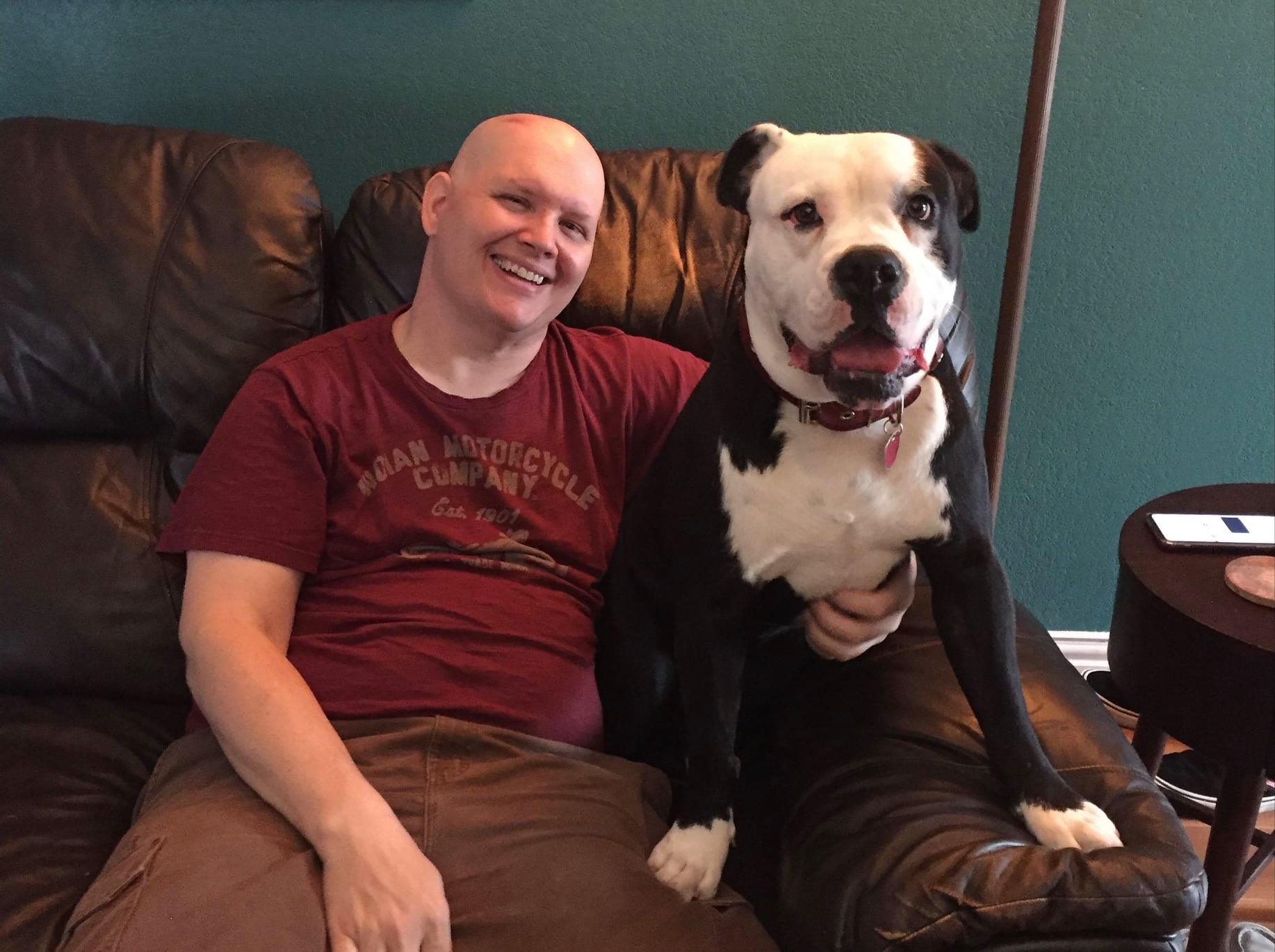 Man sitting on a couch smiling with a large black and white dog beside him, in a room with teal walls.