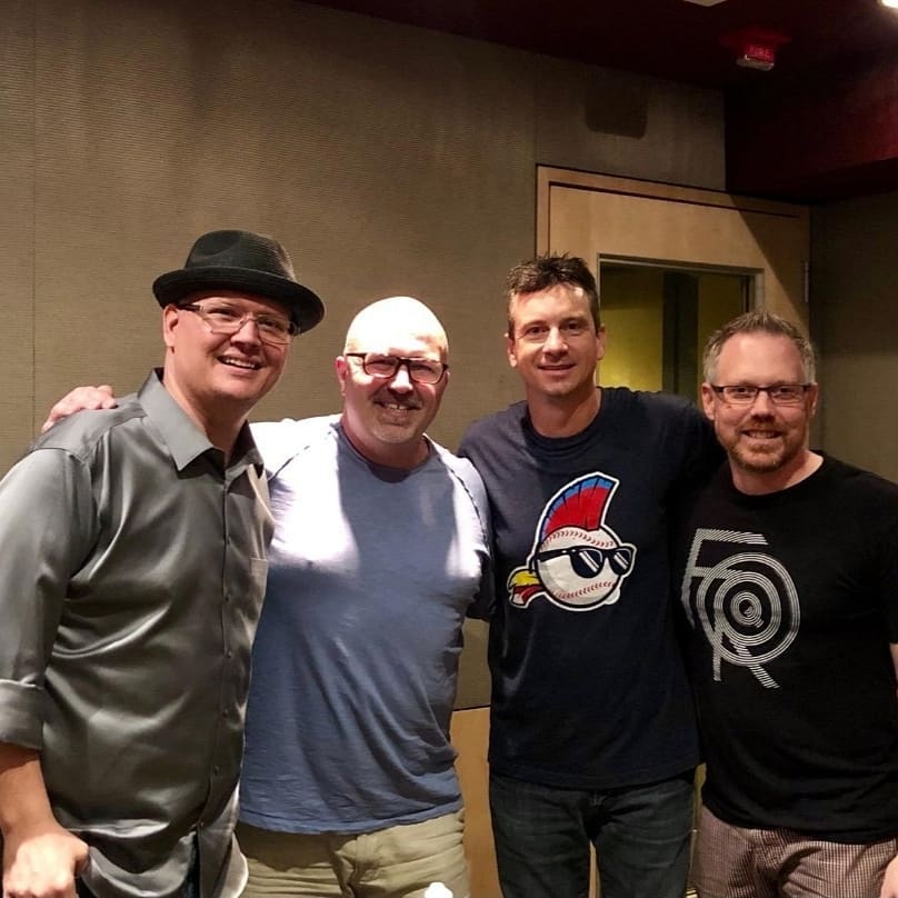 Four men standing together indoors, smiling, with a plain wall and door in the background.