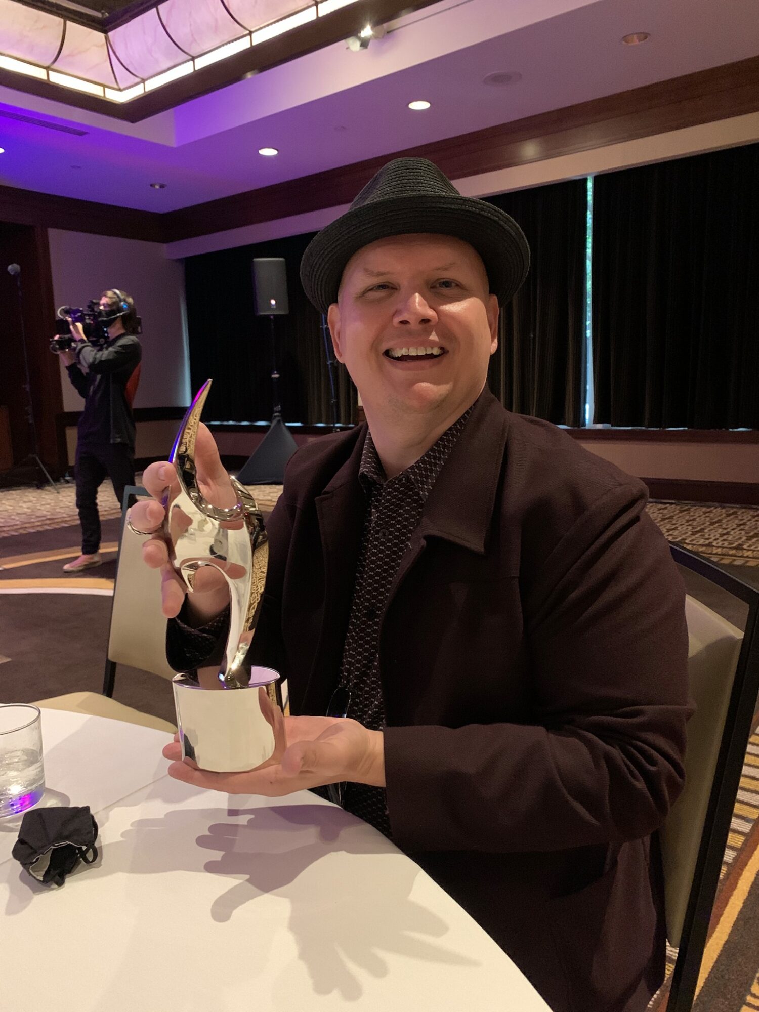 Person wearing a hat and dark jacket holding a trophy, smiling, seated at a table in a decorated event space.