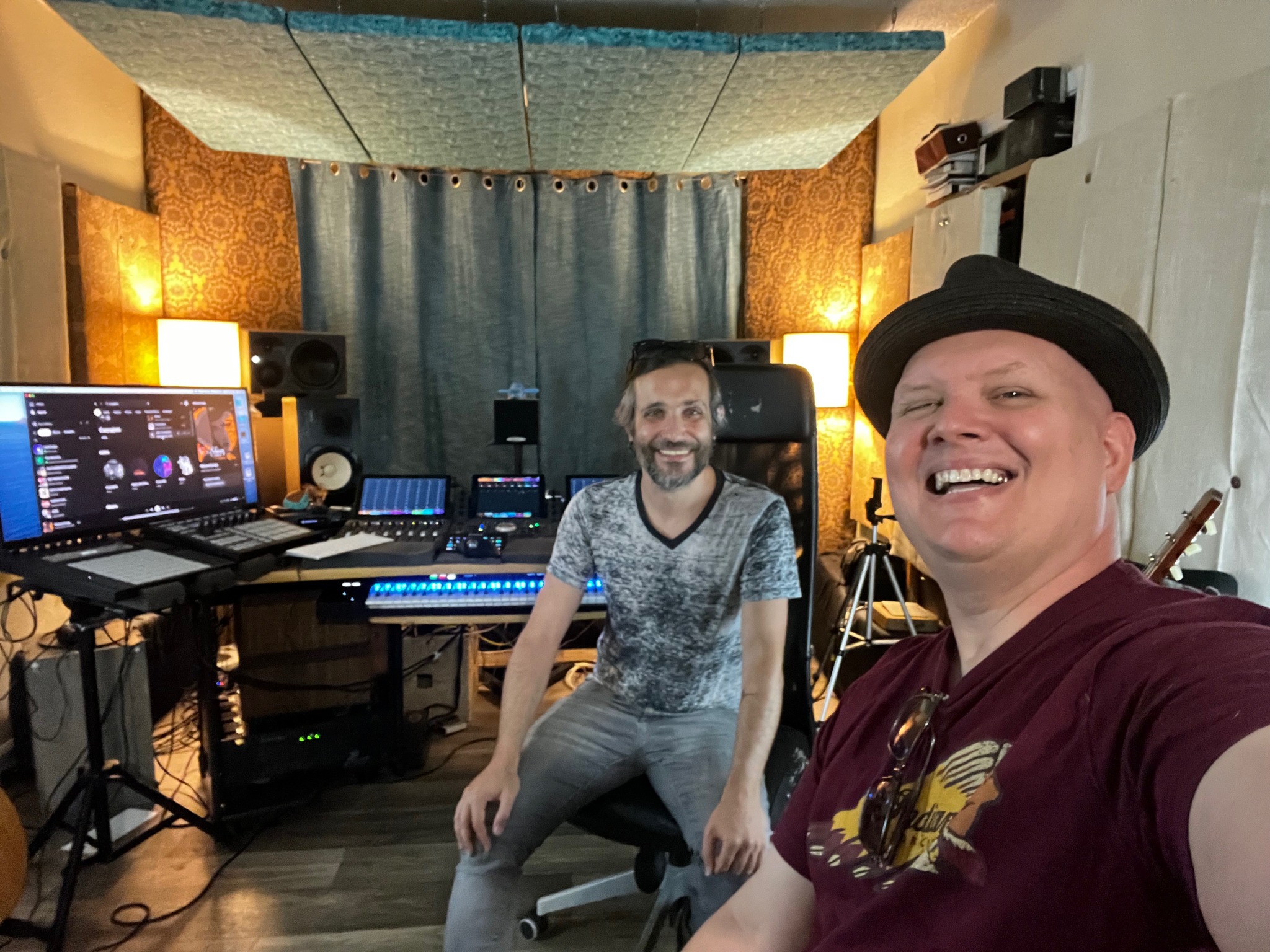 Two men smiling in a music studio with equipment, speakers, and a computer screen, one wearing a hat and the other in a t-shirt.