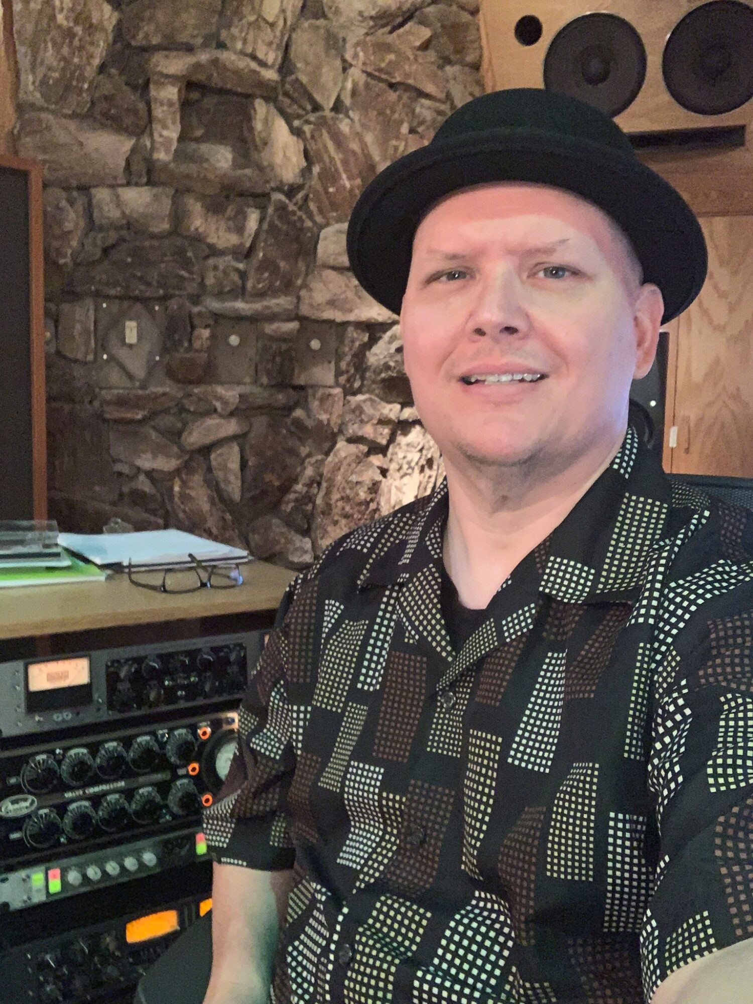 Person wearing a black hat and patterned shirt, smiling, in a room with stone wall and audio equipment.