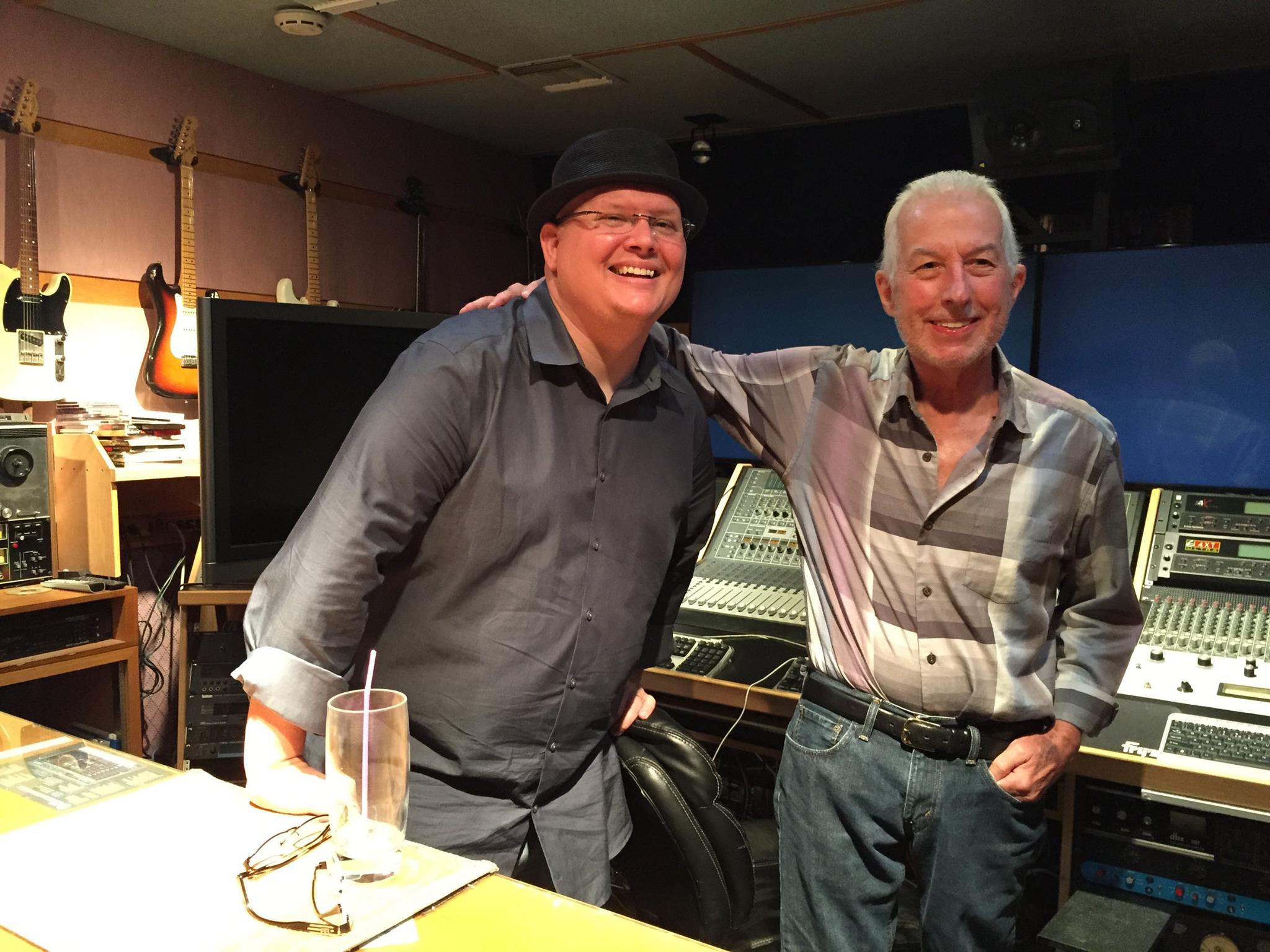 Two men smiling and standing in a music studio with equipment and guitars in the background.
