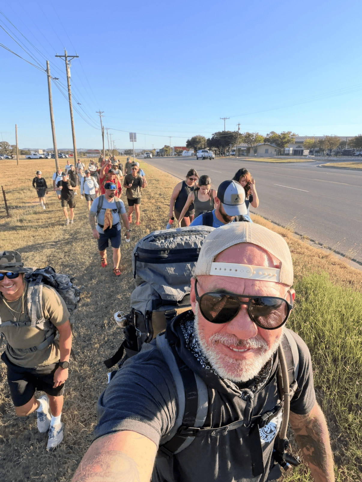 Man with sunglasses and cap taking selfie on sidewalk with group walking behind him, road and power lines in background.