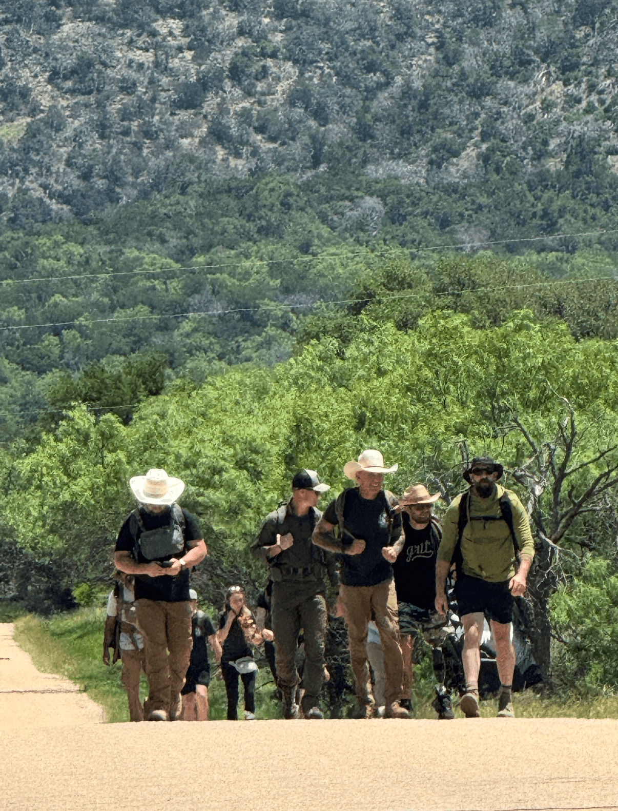 Group of people hiking on a trail with trees and mountains in the background, some wearing hats and backpacks.
