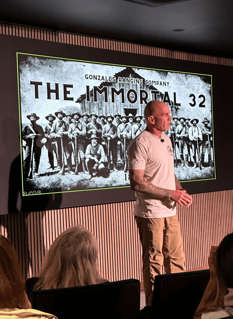 Man standing in front of a large screen displaying a historical photo of a group of people, with text about Gonzales Booting Company and The Immortal 32.