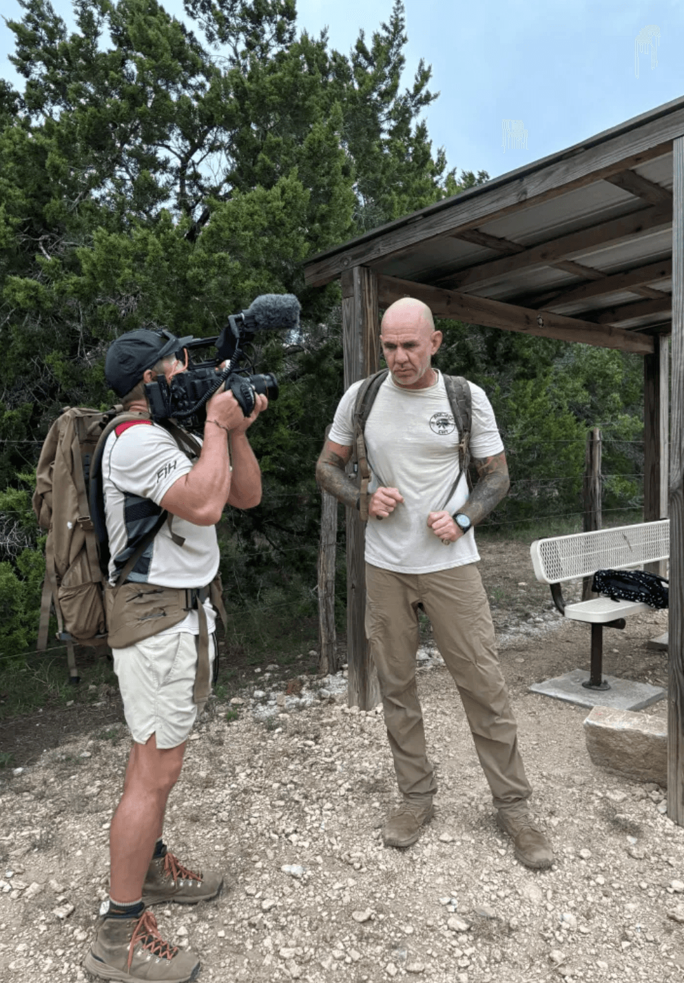 Two men outdoors, one filming the other, with trees and a wooden shelter in the background.