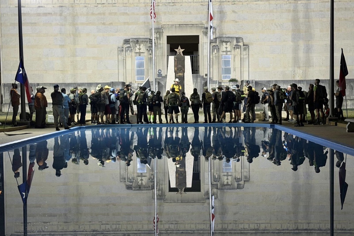 Group of people gathered in front of a large building with flags, reflected in a pool of water.