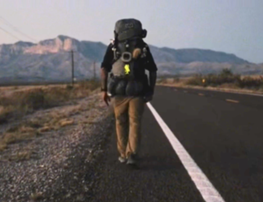 Person walking on road with large backpack, mountains in background, clear sky, white line on road, desert landscape.
