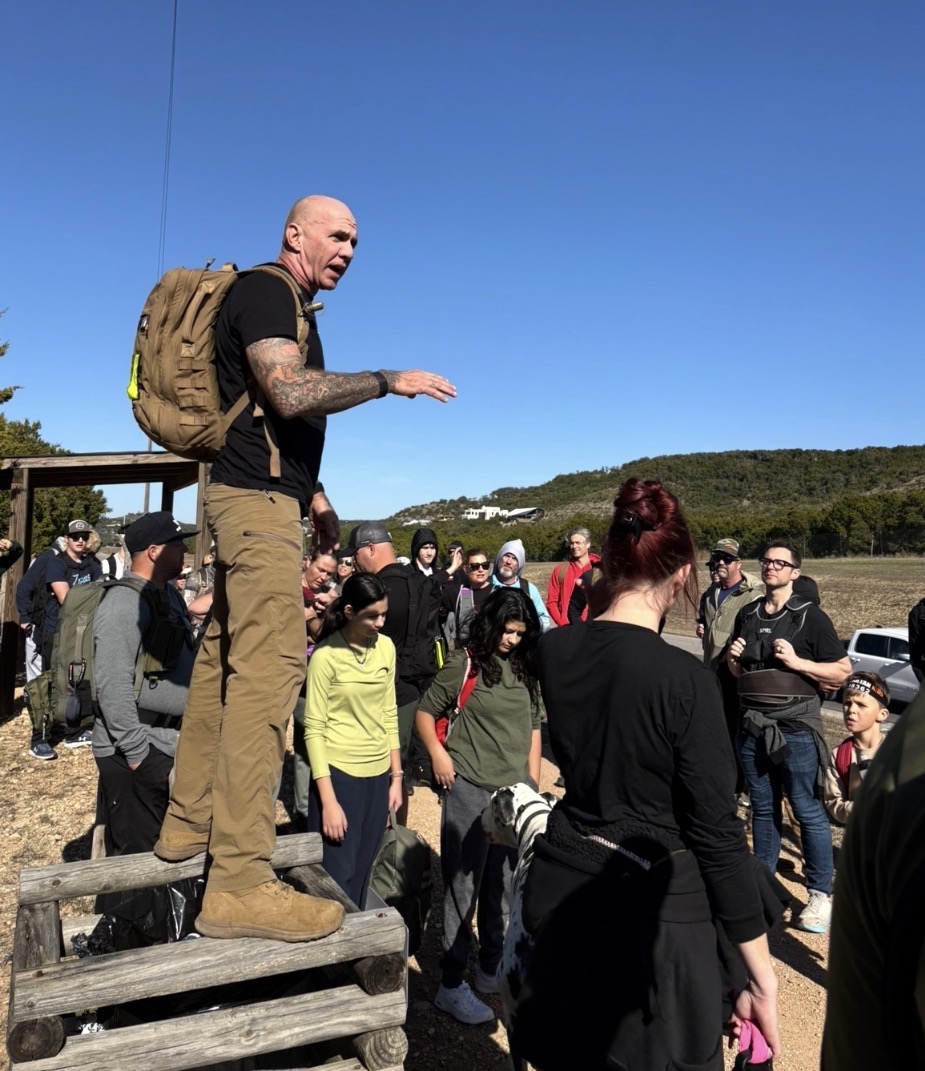 Man standing on a wooden platform addressing a group outdoors under a clear blue sky.