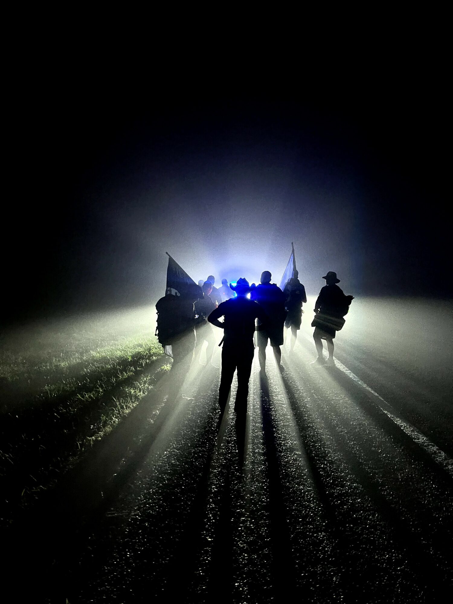 Group of people walking on a foggy road at night with bright light behind them, creating long shadows.