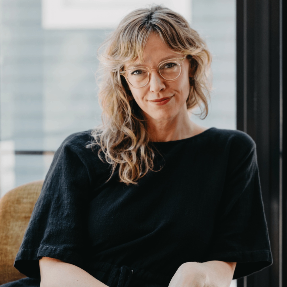 Woman with wavy blonde hair and glasses sitting indoors near a window, wearing a black top.