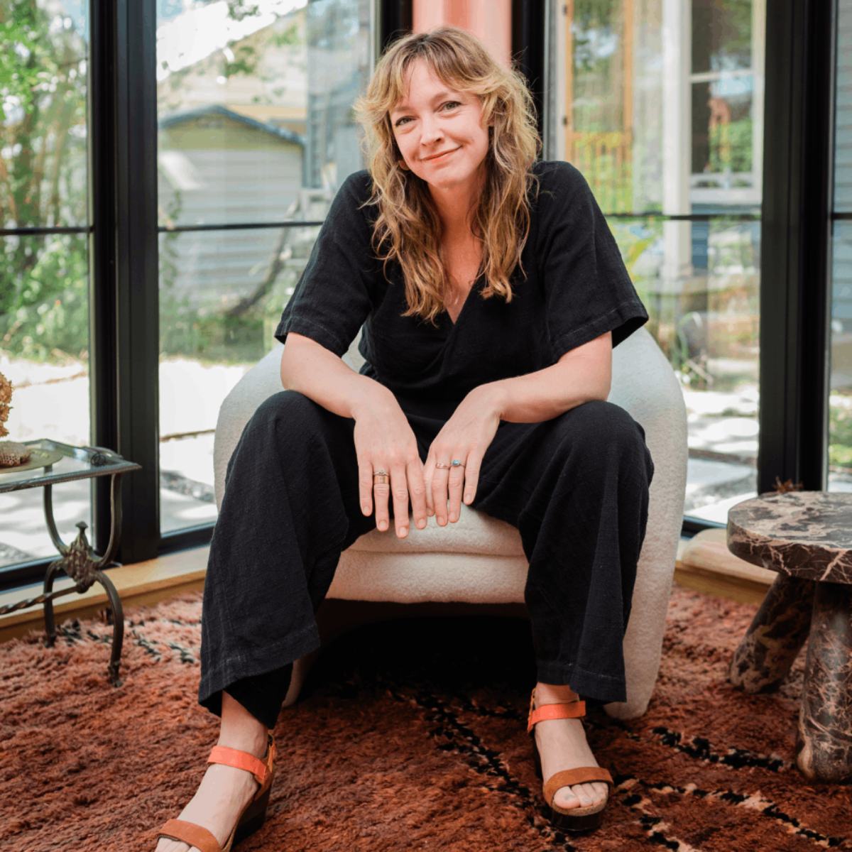 Woman sitting on a chair indoors, smiling, with large windows and greenery outside behind her.