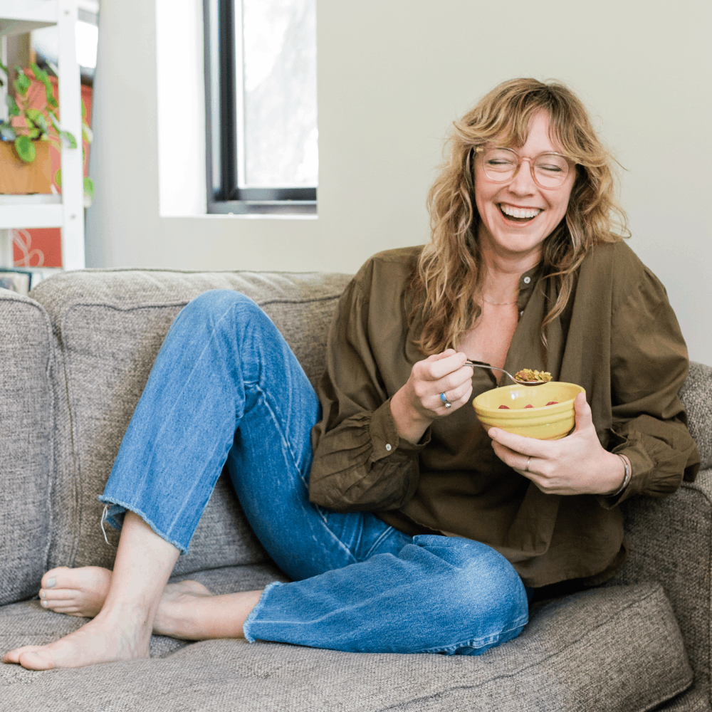 Woman with curly hair and glasses sitting on a sofa, smiling, holding a bowl of food, wearing a brown jacket and blue jeans.