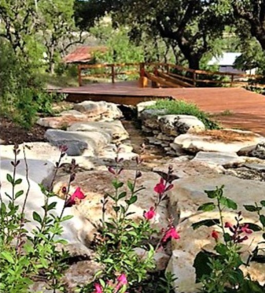 Wooden bridge over a rocky stream with plants and trees, background includes a house and greenery.