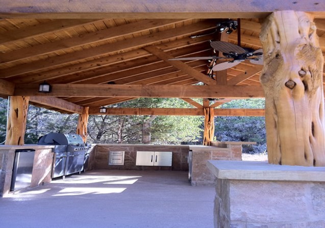 Open outdoor pavilion with wooden beams and large tree trunk columns, kitchen area with counters, and trees in background.