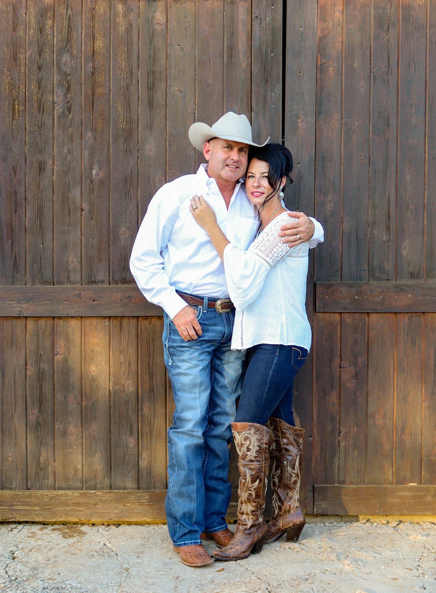 A man in a cowboy hat and woman in a white top stand close together, smiling, in front of a wooden fence.