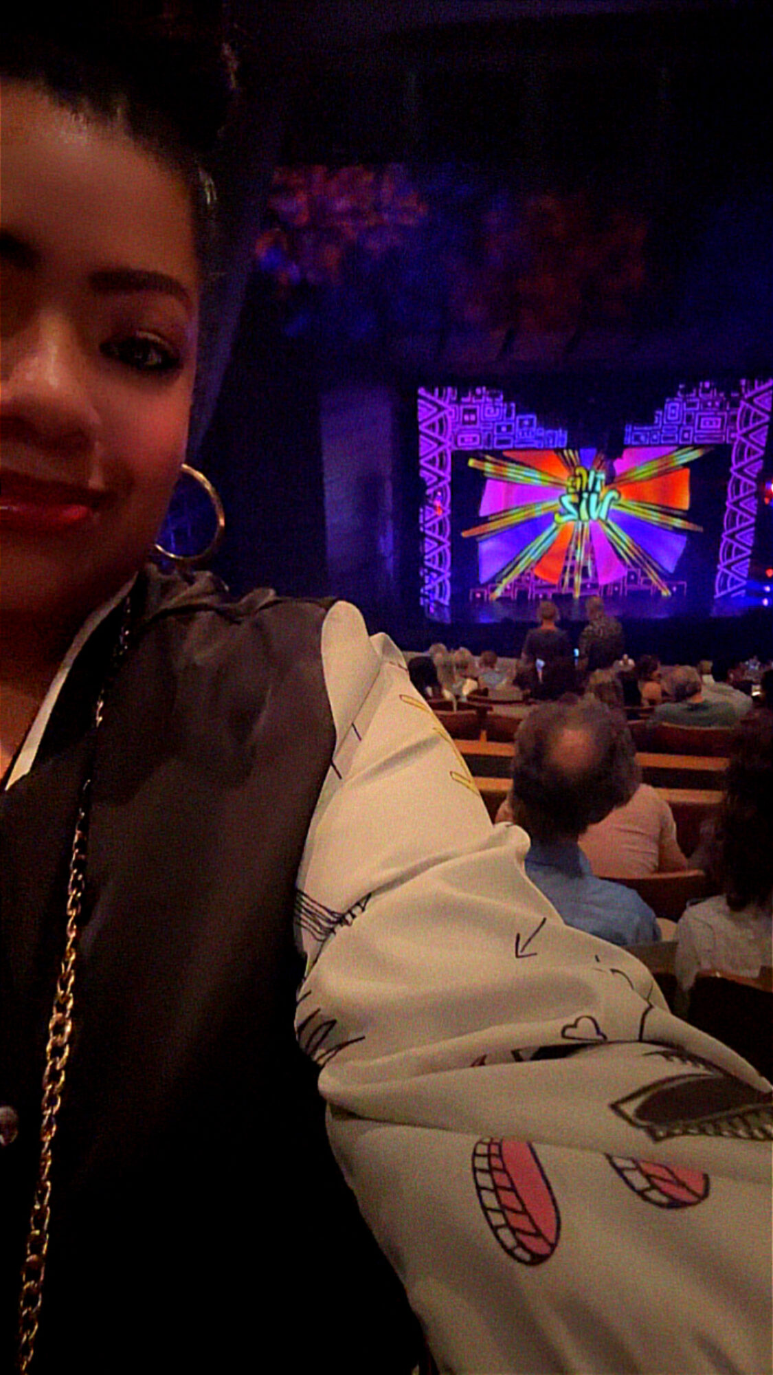 Partially visible person taking selfie at concert with colorful stage lights and audience in background.