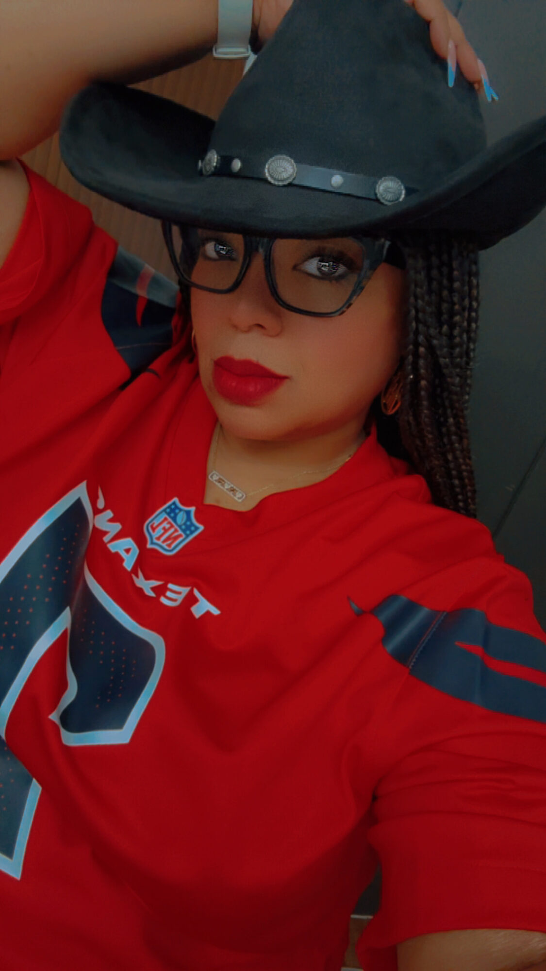 Person wearing glasses, red sports jersey, and a large black hat, posing with hand on hat, against dark background.