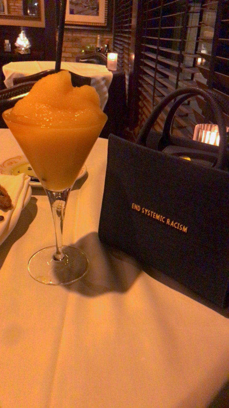 A yellow frozen drink in a tall glass on a table with a black bag nearby, in a dimly lit restaurant.