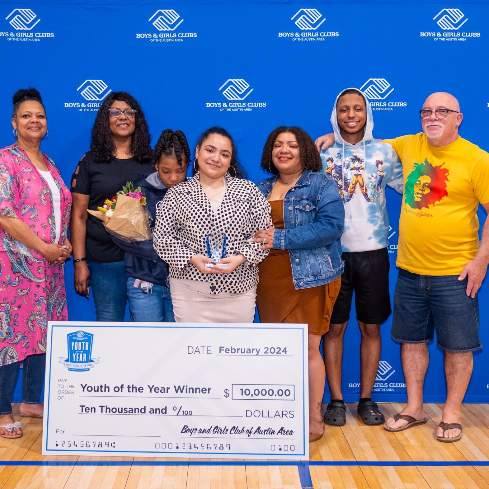 Group of seven people standing indoors, holding a large check for youth award, smiling, with a blue backdrop. Word count: 20