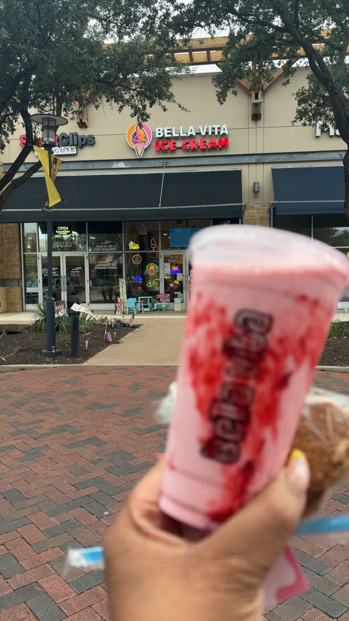 Person holding a pink ice cream cone in front of a storefront with trees and a brick sidewalk.