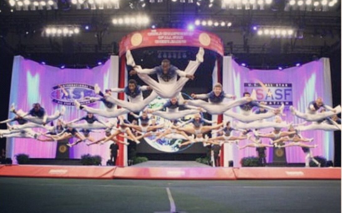 Group of gymnasts performing a synchronized routine on a stage with colorful lighting and a backdrop, mid-air with extended limbs.
