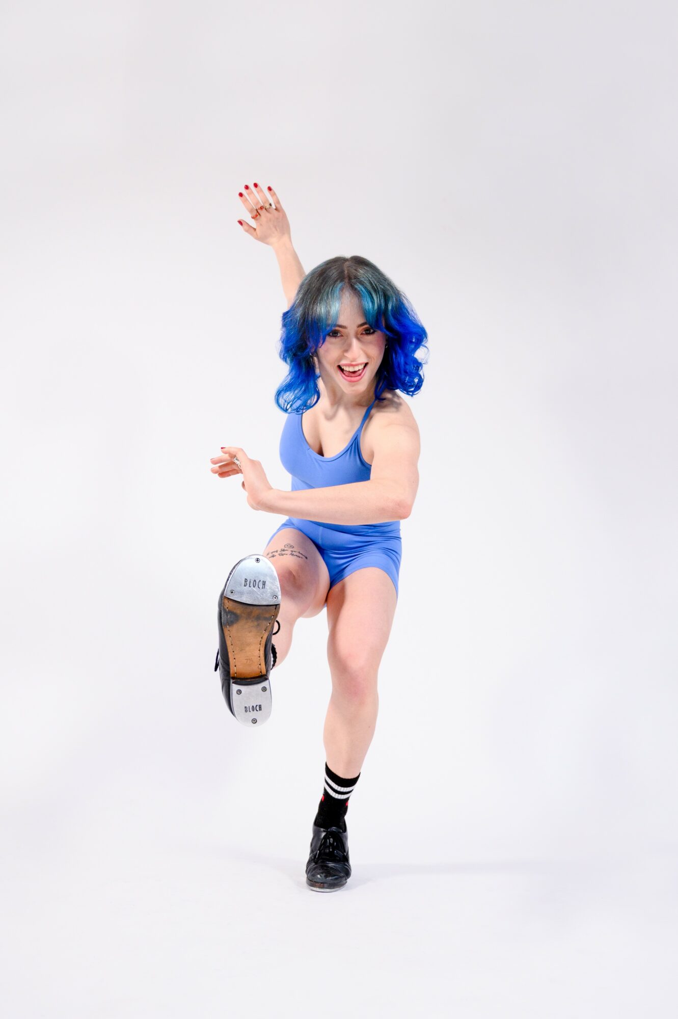 Young woman with blue hair jumping with one leg raised and arms outstretched, smiling, against a plain white background.