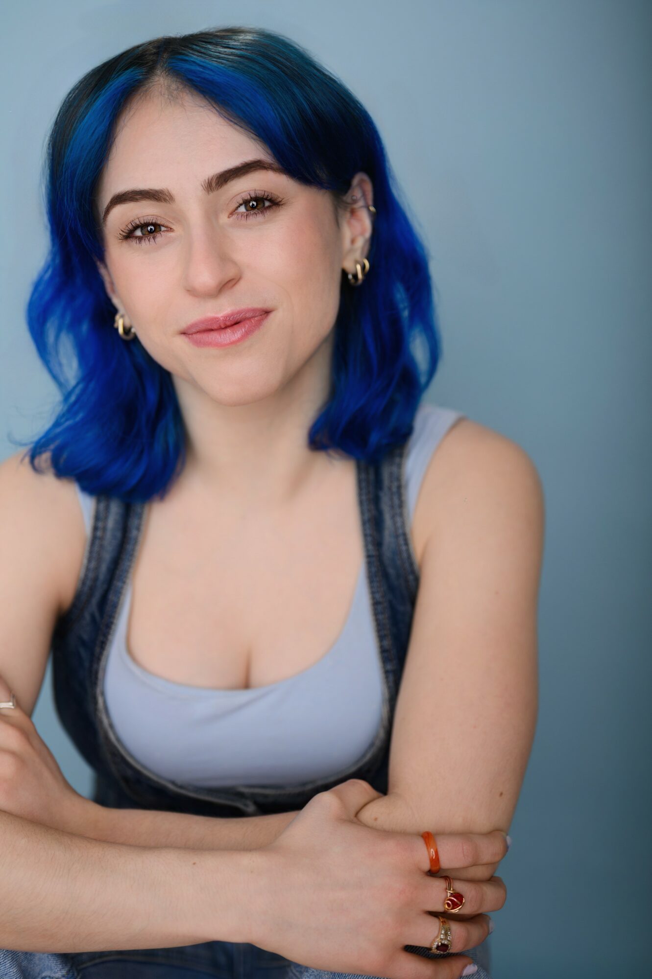 Young woman with blue hair, smiling, wearing a light gray top and earrings, against a blue background.