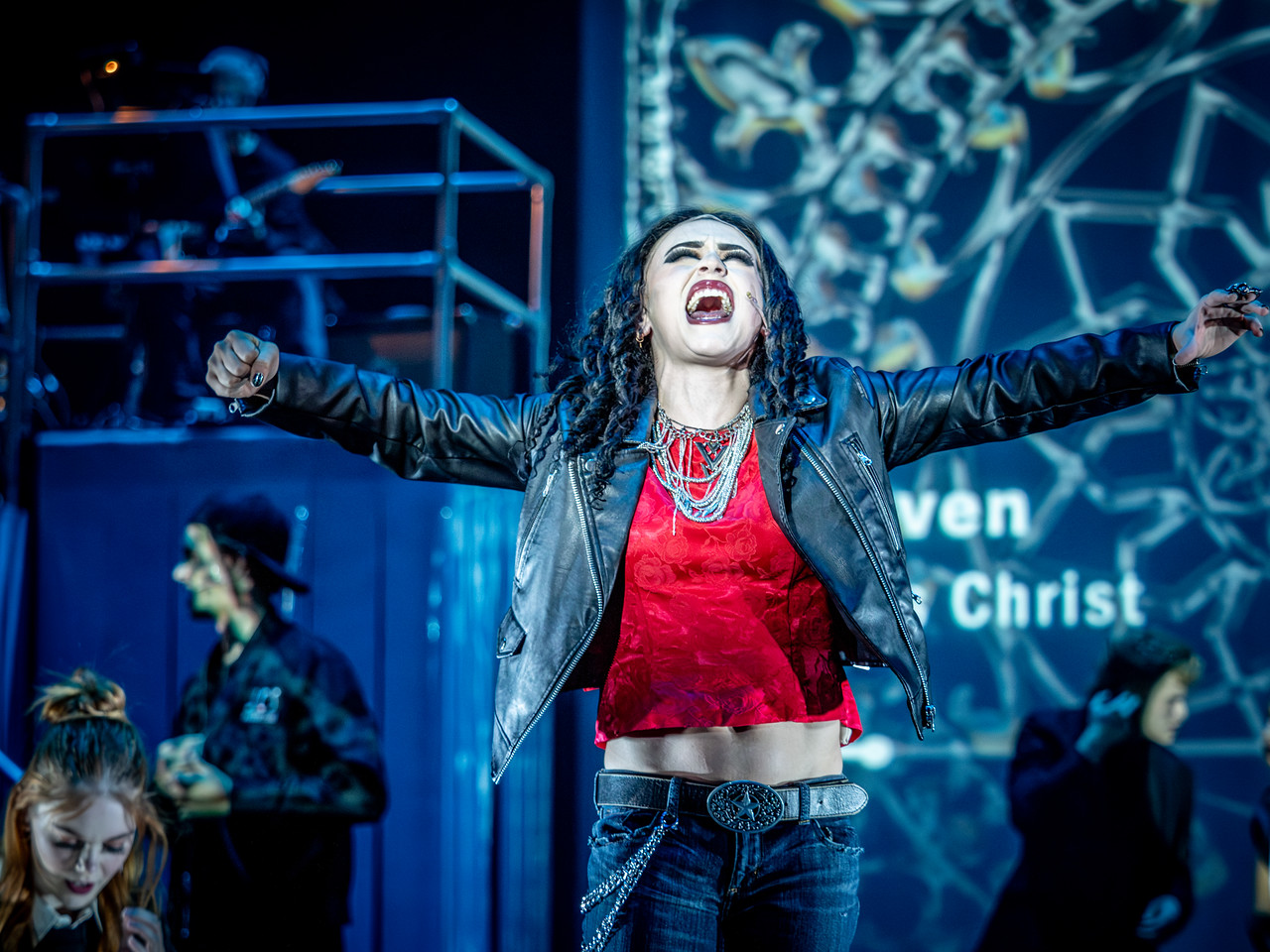 Woman with arms outstretched singing on stage, wearing a leather jacket and red top, with a dark background and stage lights.