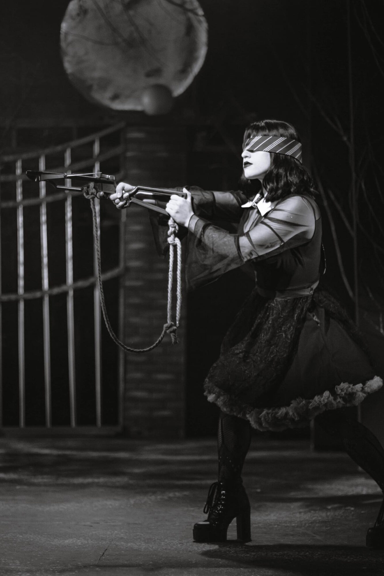 Performer in costume holding a rifle prop on stage, with a moon backdrop and railing, in black and white.