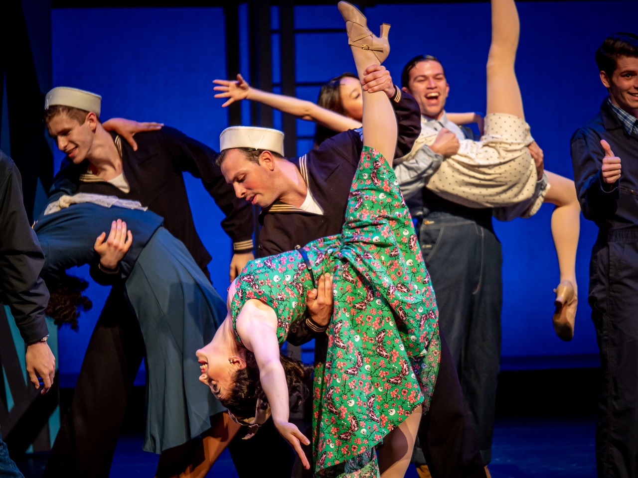 Group of performers on stage, one person in a colorful dress is upside down, others are smiling and posing.