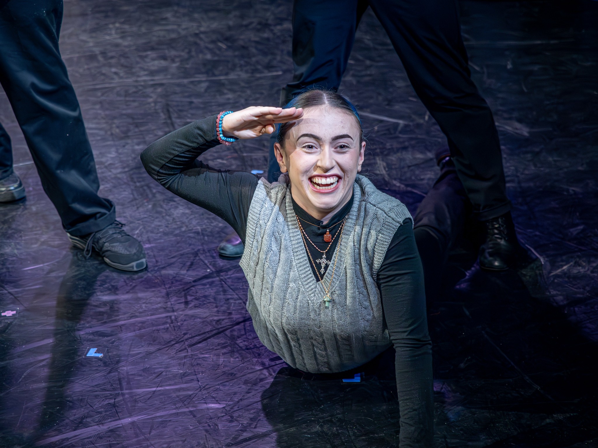 Smiling woman on stage, saluting with her right hand, surrounded by people, dark stage background