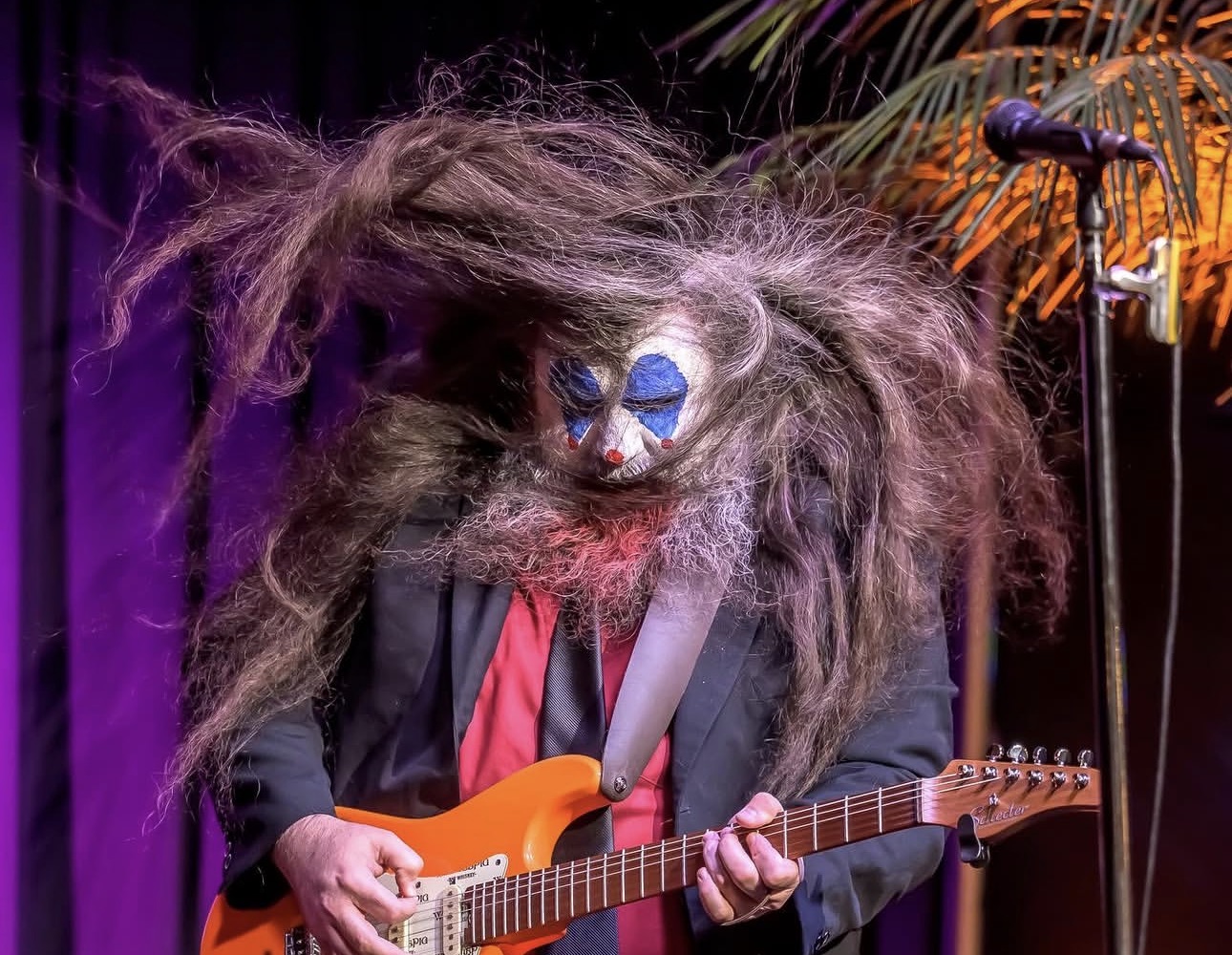 Person in a mask with wild hair playing an orange electric guitar on stage with purple lighting.