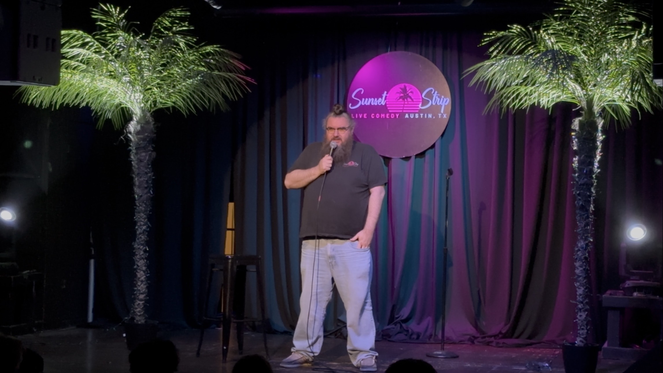 Man standing on stage with microphone, curtains, and palm trees in background, performing comedy or speech.
