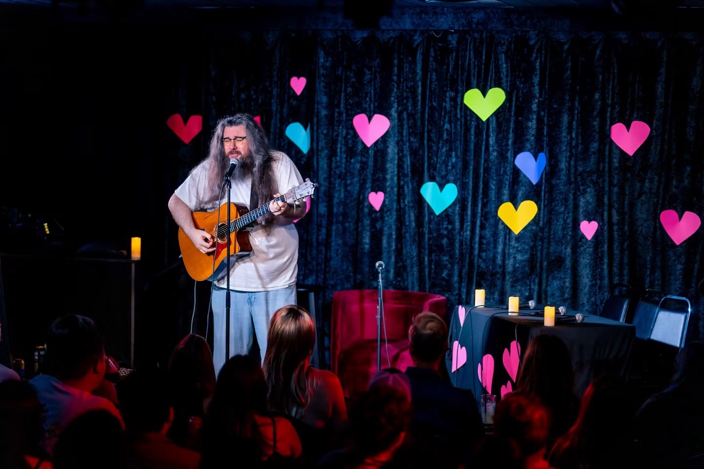 Person with long hair playing guitar on stage with heart-shaped decorations, audience watching, dark background, candles on table