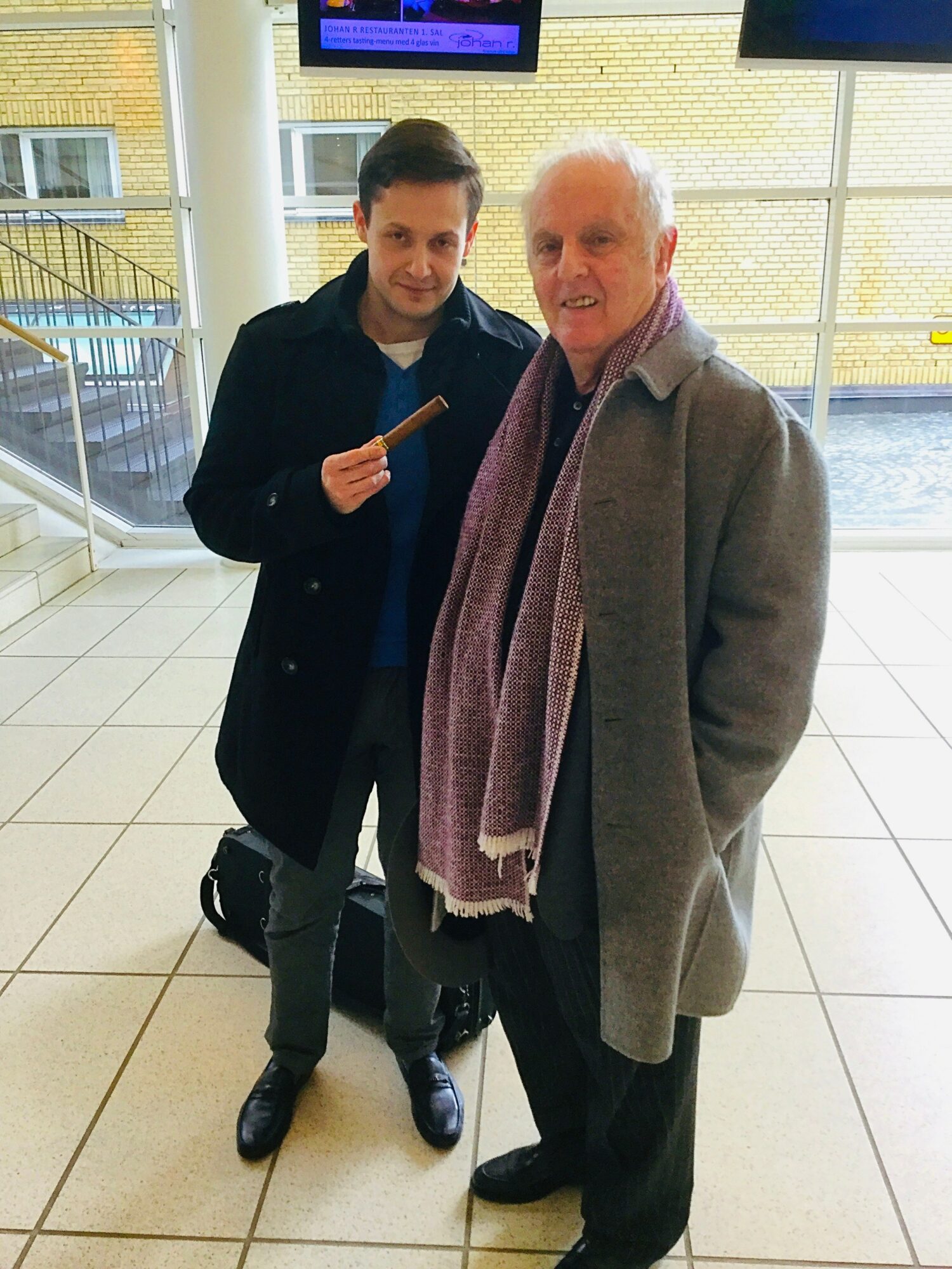 Two men standing indoors, one holding a small object, with large windows and a digital screen behind them.
