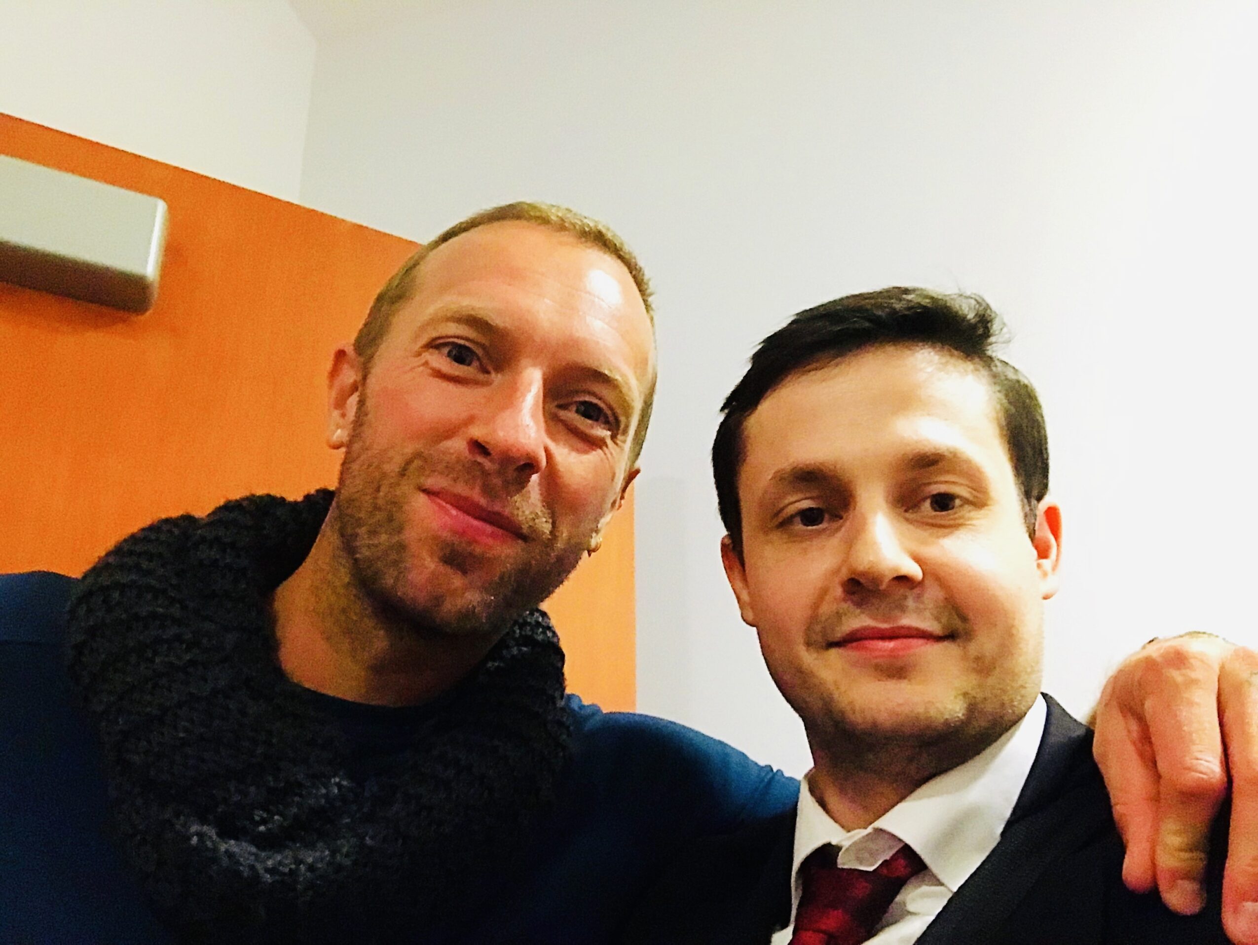 Two men smiling, one with arm around the other, in an indoor setting with a white wall and orange panel behind.