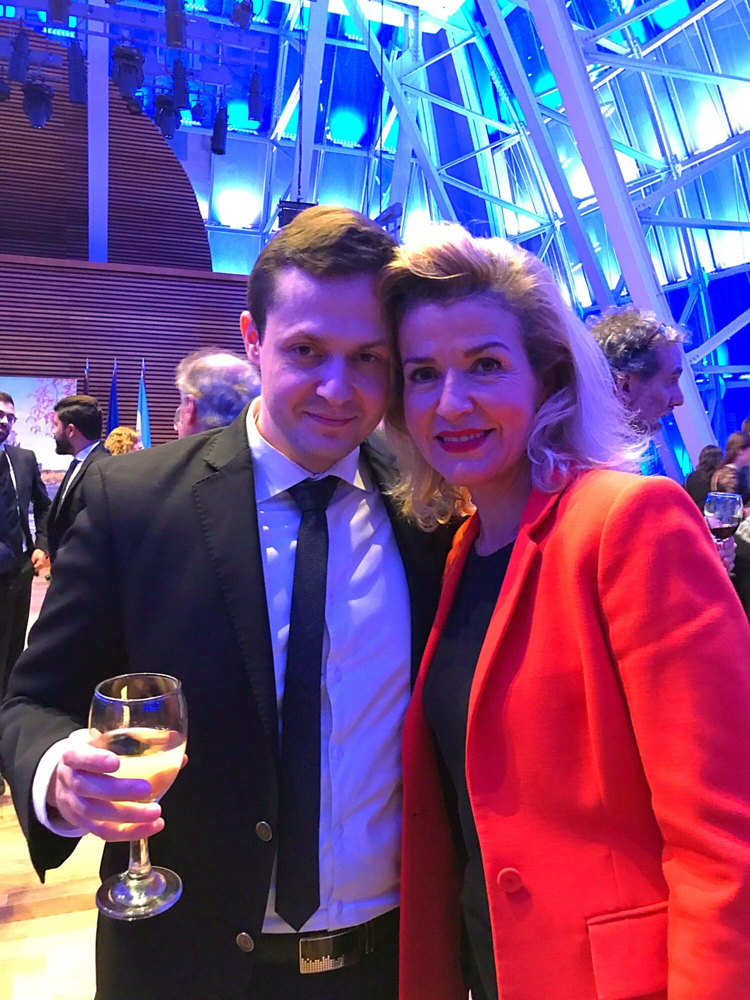 Two people, a man in a suit and a woman in a red blazer, pose together at an event with blue lighting.