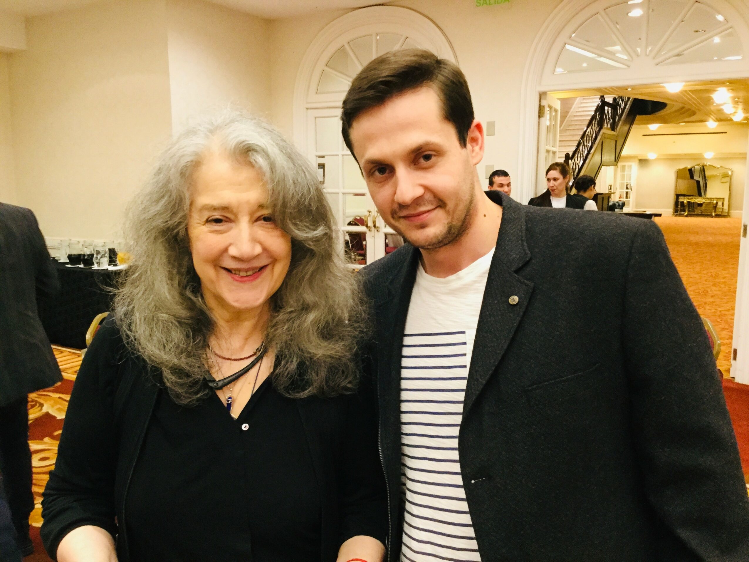 A smiling woman with long gray hair and a young man with short dark hair pose together indoors, with warm lighting and a piano in the background.