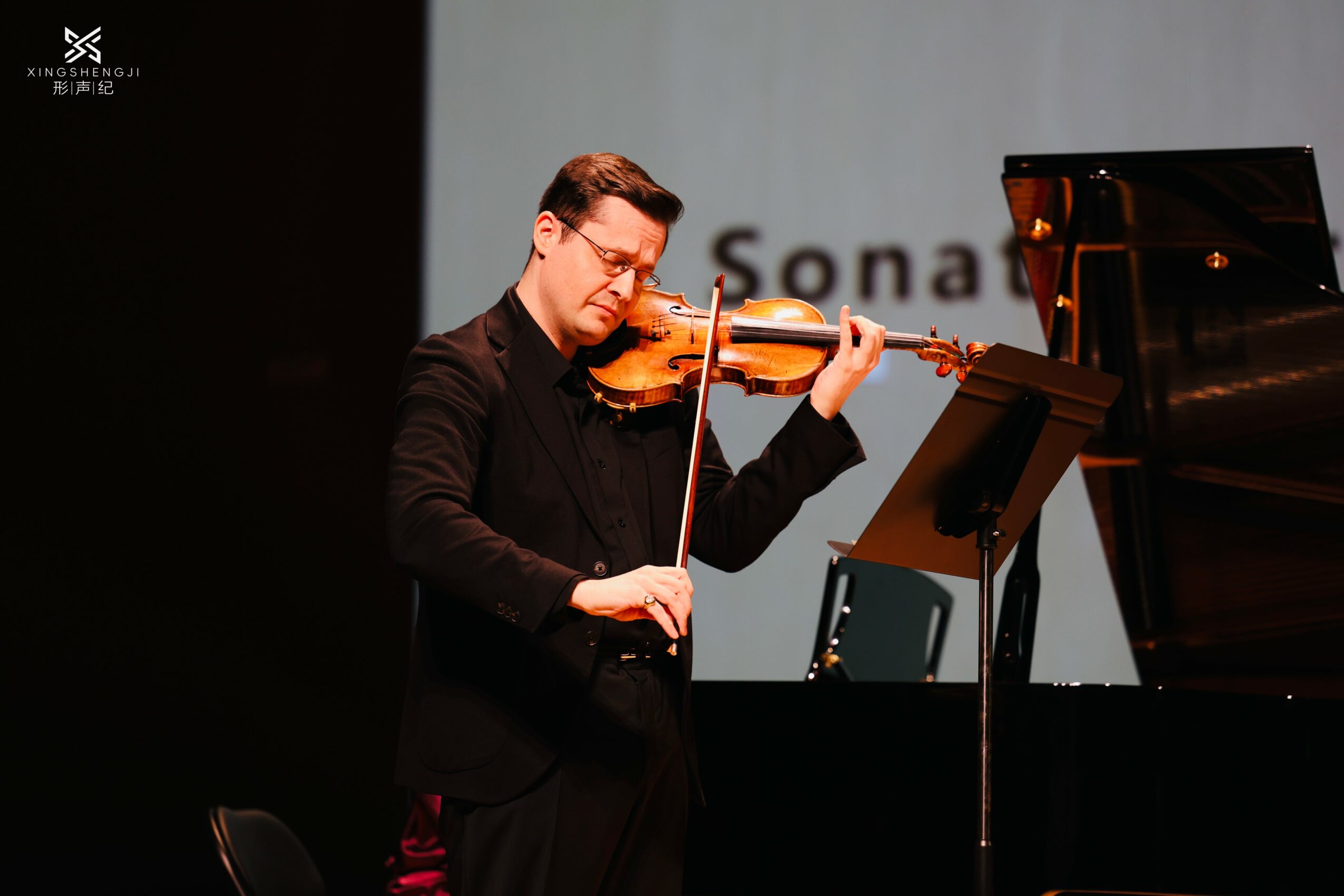 A man playing a violin in front of a piano, with a music stand, on stage with a gray background.