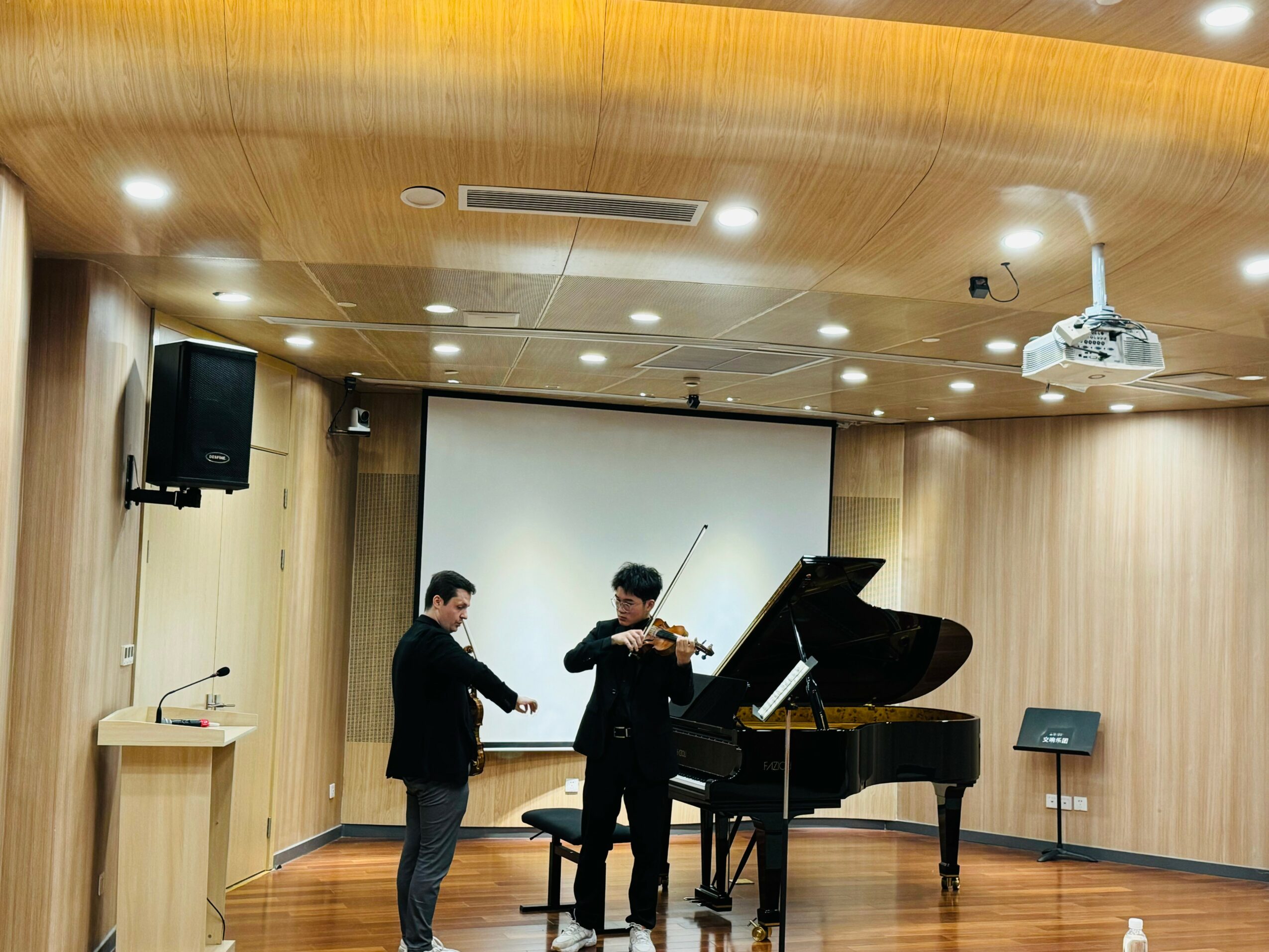 Three people in a music room, one playing the violin, another standing nearby, with a grand piano and a projection screen behind them.