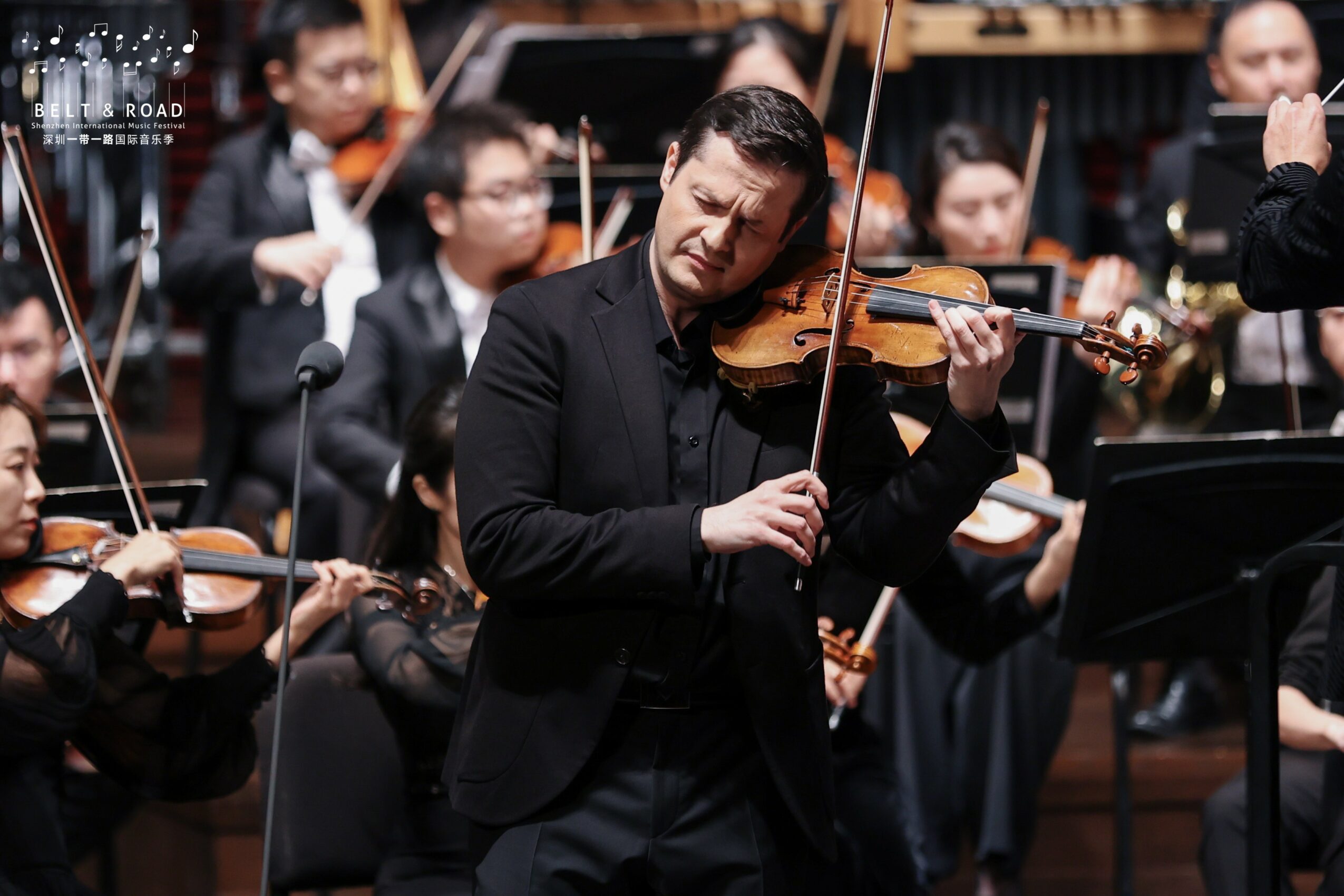 Orchestra with a violinist playing passionately, surrounded by other musicians in formal attire, in a concert hall setting.