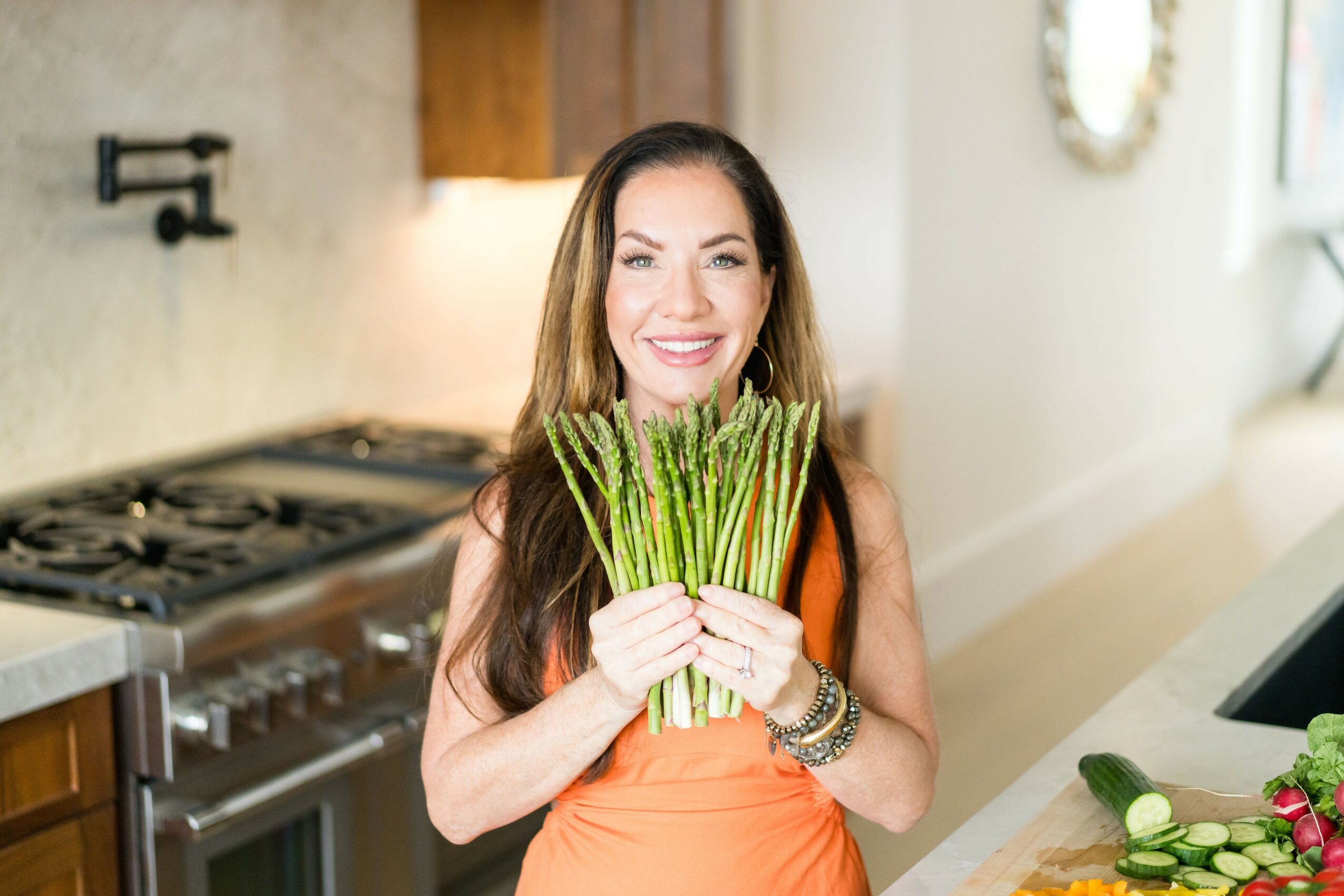 Woman with long brown hair smiling, holding a bunch of green asparagus in a kitchen.