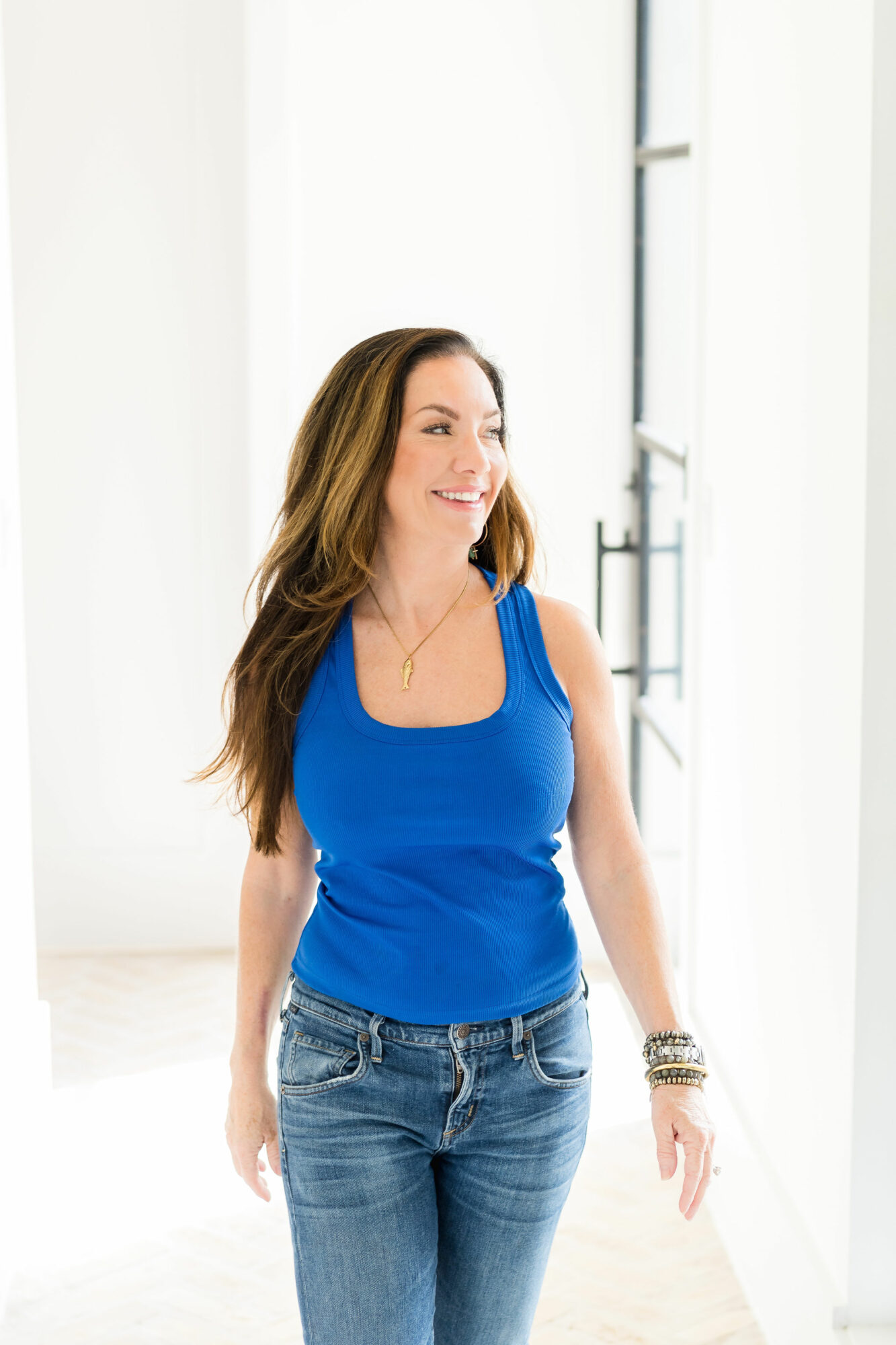 Woman with long hair wearing a blue tank top and jeans, smiling indoors near a window.