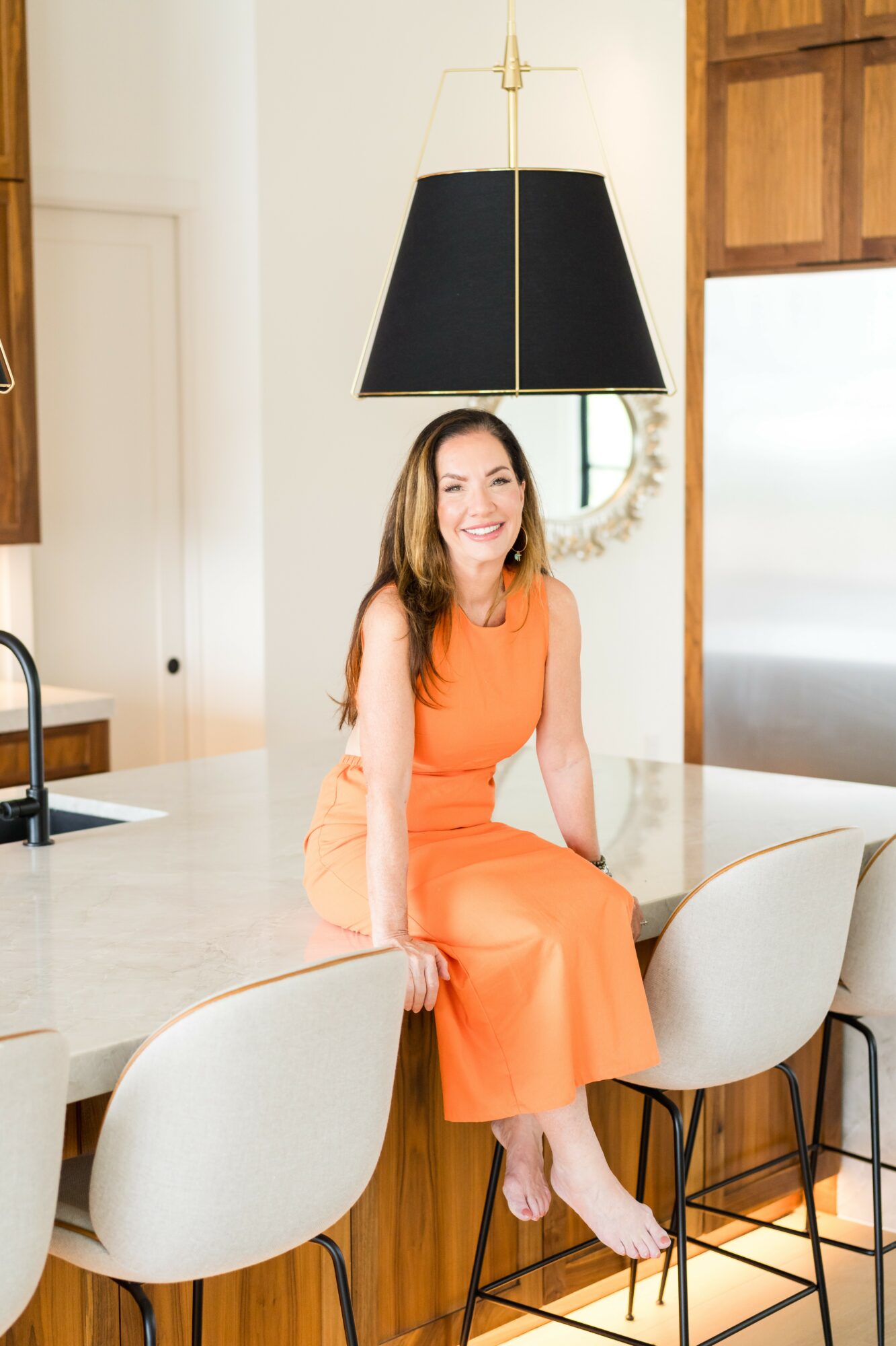 Woman in orange dress sitting on kitchen counter, smiling, with black lampshade hanging above, wooden cabinets, and window in background.
