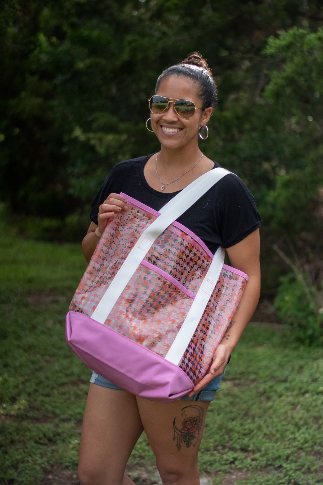 Smiling woman with sunglasses holding a large pink and patterned tote bag outdoors, with trees in the background.
