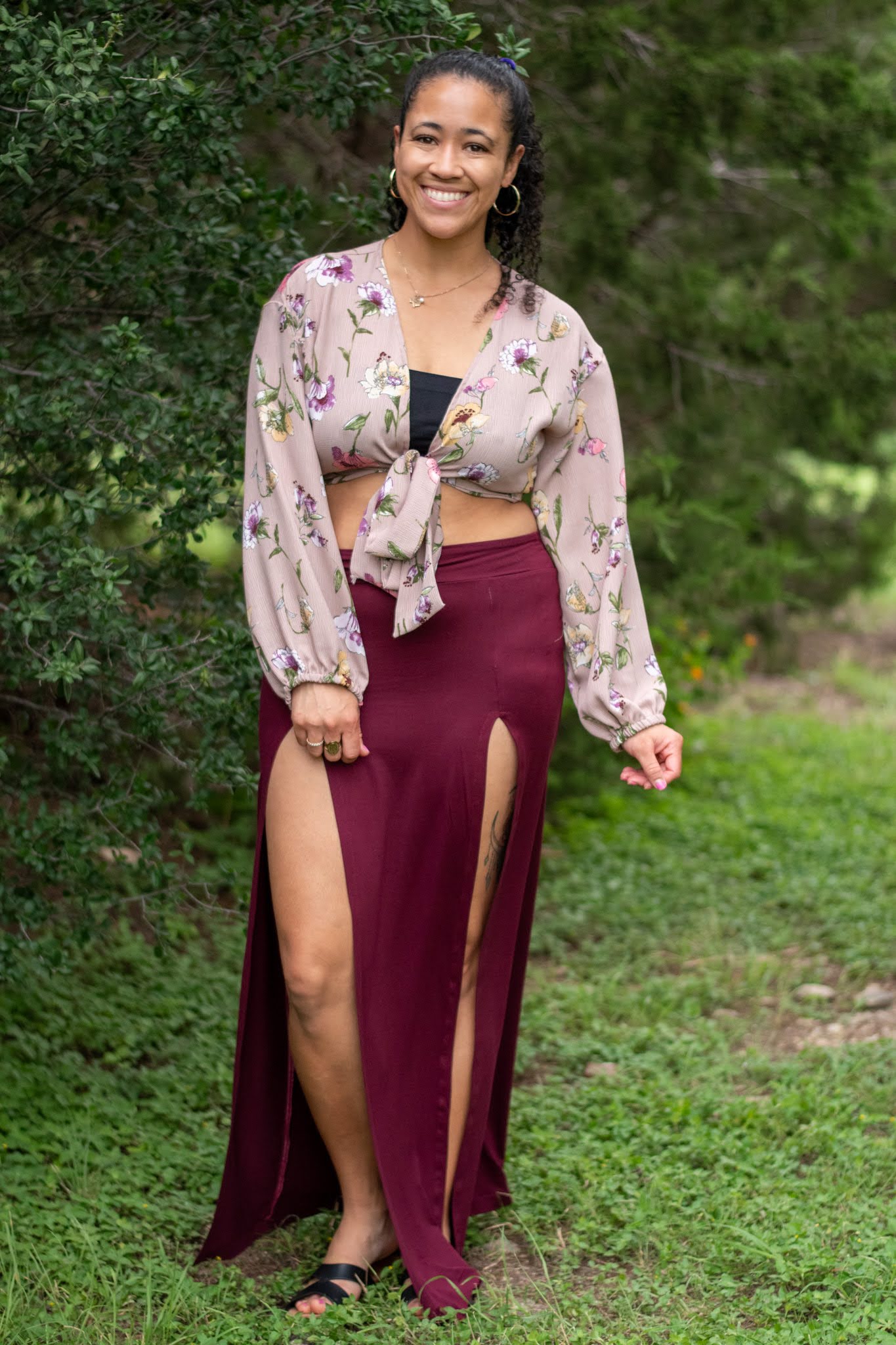Smiling woman standing outdoors on grass, wearing a floral blouse and a long maroon skirt with high slits.
