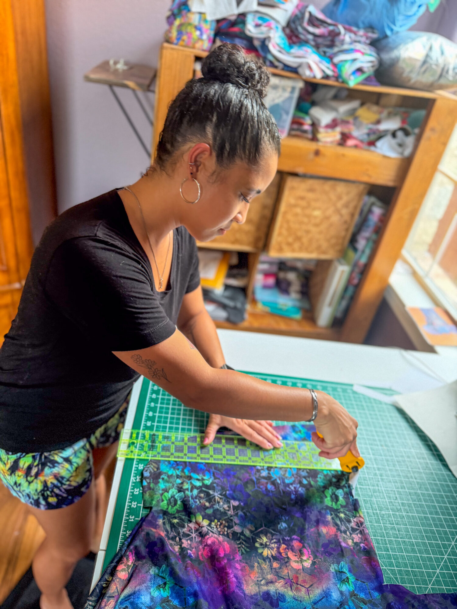 Woman working on fabric at a sewing table in a room with shelves and a window.