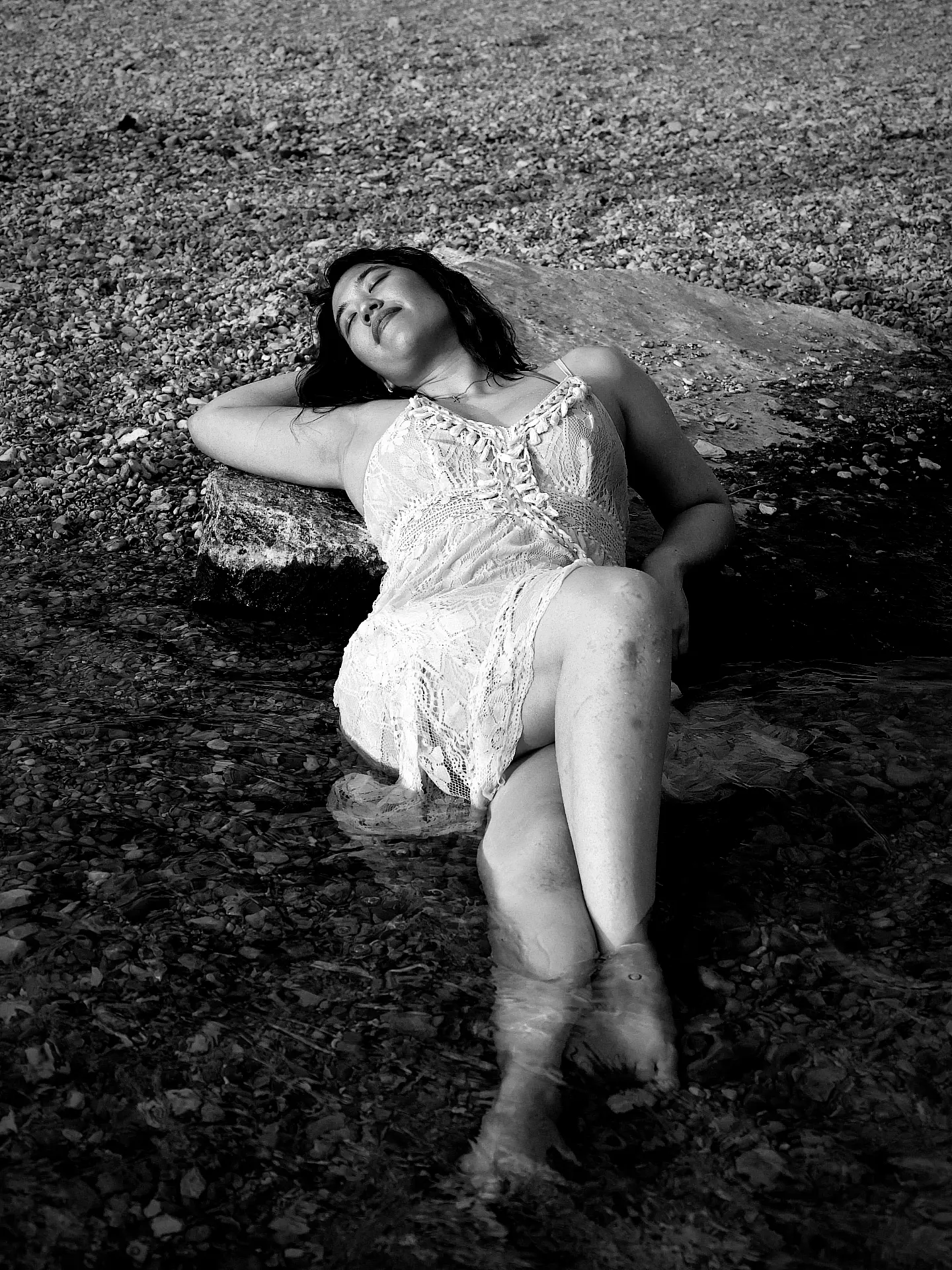 Woman in a dress lying on a rock with water around her, eyes closed, head resting on her arm, black and white photo.