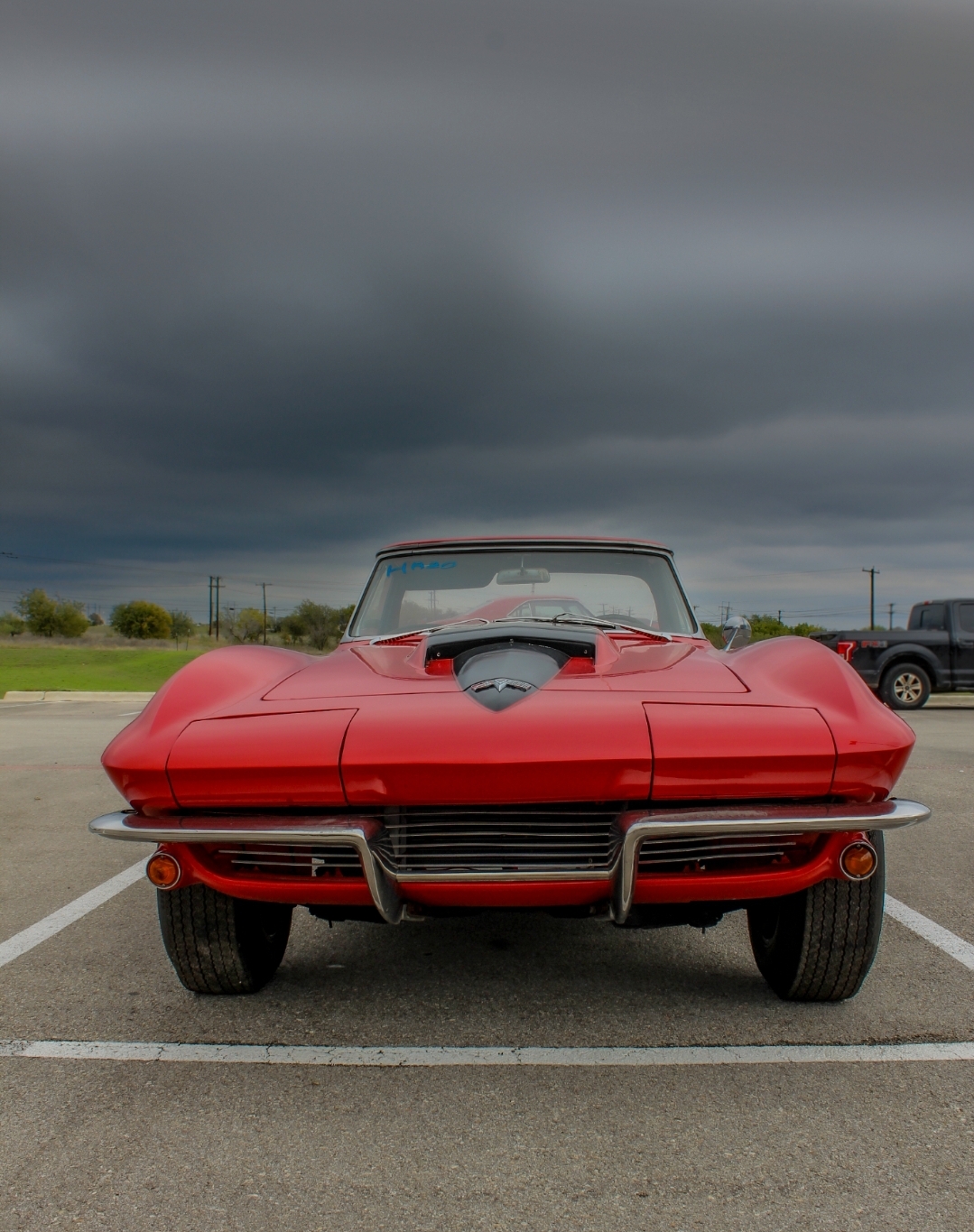 Red classic car parked in an empty parking lot under dark, cloudy sky, facing forward with visible front grille and hood scoop.